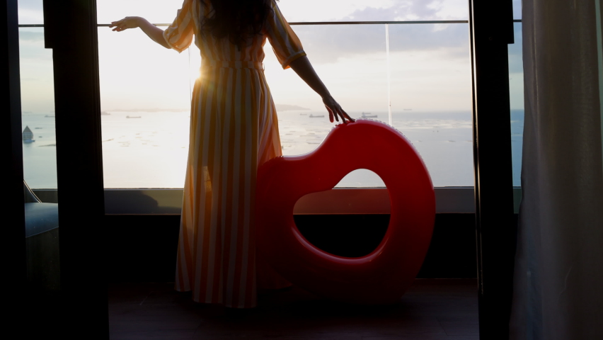 Beautiful woman from back in yellow dress and hat holding rubber swim tube of heart shape on open air window terrace with sea view and orange sunset sky calmness tranquil relaxing sunlight summer.  