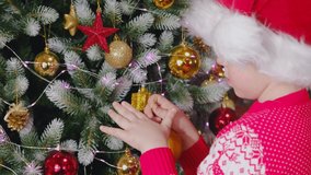 Merry Christmas kids. A child removes a golden ball from a Christmas tree, wearing a Santa's New Year's hat. Christmas and New Year. Video clip