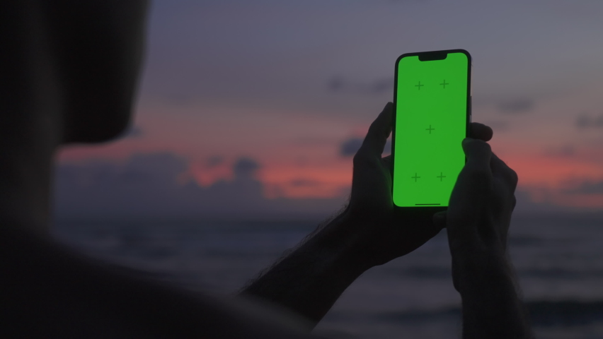 Silhouette man holds vertical mobile phone one hand and swiping touch screen. Use green screen for copy space closeup. Chroma key mock-up on smartphone. Film pixel grain texture.  Royalty-Free Stock Footage #1096371801