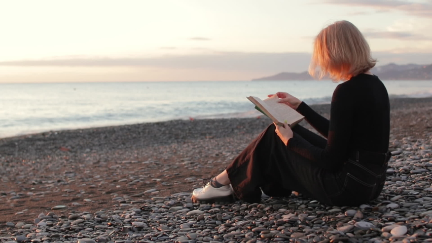 Young woman reading a book by the sea at sunset. Woman relaxing on beach. Royalty-Free Stock Footage #1096373325