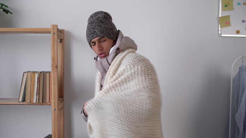 Young man warmly dressed and wrapped in a blanket feeling cold freezing at home in winter. No heating