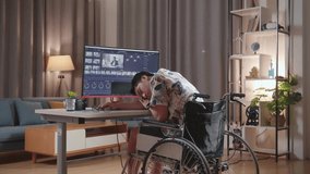 Back View Of Asian Man In Wheelchair Sleeping While Editing And Color Grading The Video By A Desktop Next To The Camera At Home.