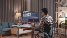 Back View Of Asian Man In Wheelchair Holding His Chin And Thinking About Something While Editing The Video And Color Grading By A Desktop Next To The Camera At Home