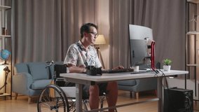Asian Man In Wheelchair Thinking About Something And Raising His Index Finger While Editing The Video By A Desktop Next To The Camera At Home.