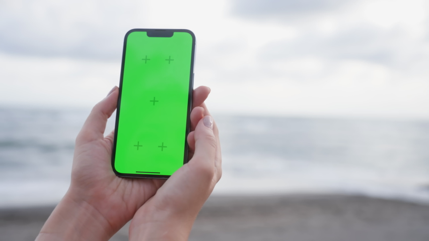 Close-up of woman's hands holding a vertical mobile phone and pressing her thumb finger into center of touch screen against background of ocean beach with people. Use green screen for copy space.  | Shutterstock HD Video #1096378279