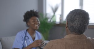 4K video footage of A happy young nurse provides medical care, helps an old mature man on a medical visit, a nursing lady gives empathy encourages a retired patient to sit on a sofa in a home hospital