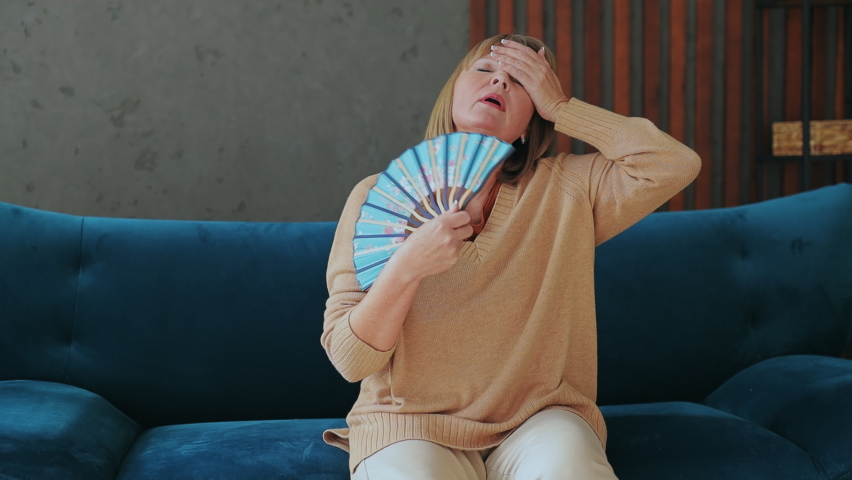 Elderly overheated woman 50s years old wears casual clothes sits on blue sofa hold in hand waving fan stay at home flat without air conditioning spend free spare time in living room indoor grey wall Royalty-Free Stock Footage #1096378985