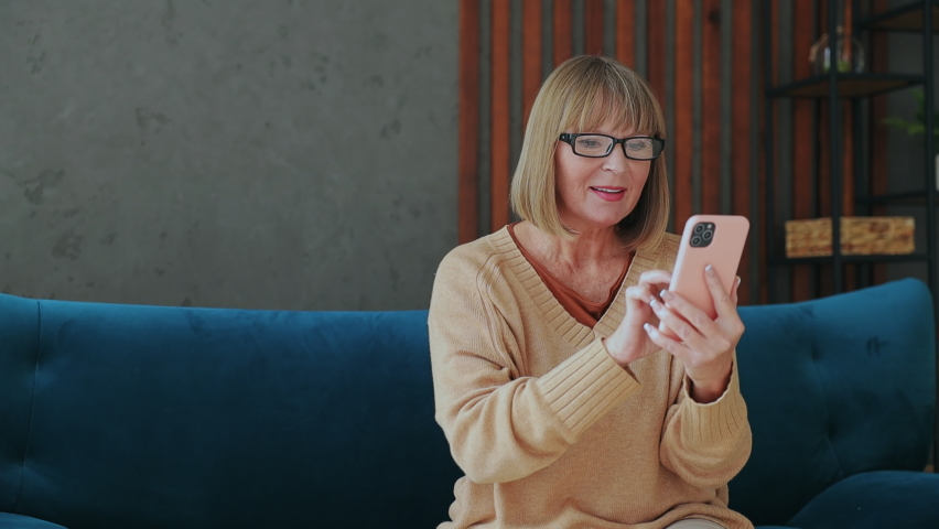 Elderly woman 50s year old wears casual clothes sits on blue sofa use mobile cell phone do winner gesture clench fist stay at home flat rest relax spend free spare time in living room indoor grey wall