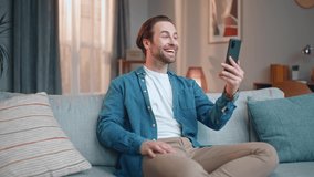 Charming happy man making video call with bestfriend using smartphone. Pretty young stylish male calling people smiling, showing thumbs up, online, sitting on sofa. Video call concept.