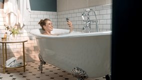 Young Caucasian pretty female smiling and video chatting on smartphone while taking relaxing bath at home. Beautiful cheerful woman in tub with bubble foam in bathroom and speaking on online call