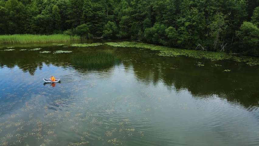 male fisherman in an orange raincoat floats on an inflatable boat on the lake in summer. drone point of view