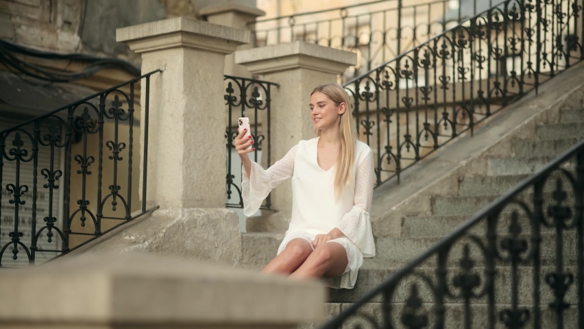Portrait smiling young woman with blonde hair sitting on stairs using mobile phone. Happy woman making facetime video calling with smartphone