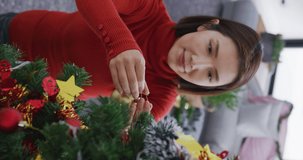 Vertical video of happy asian woman decorating christmas tree at home. Christmas, celebration, happiness, tradition and inclusivity concept.
