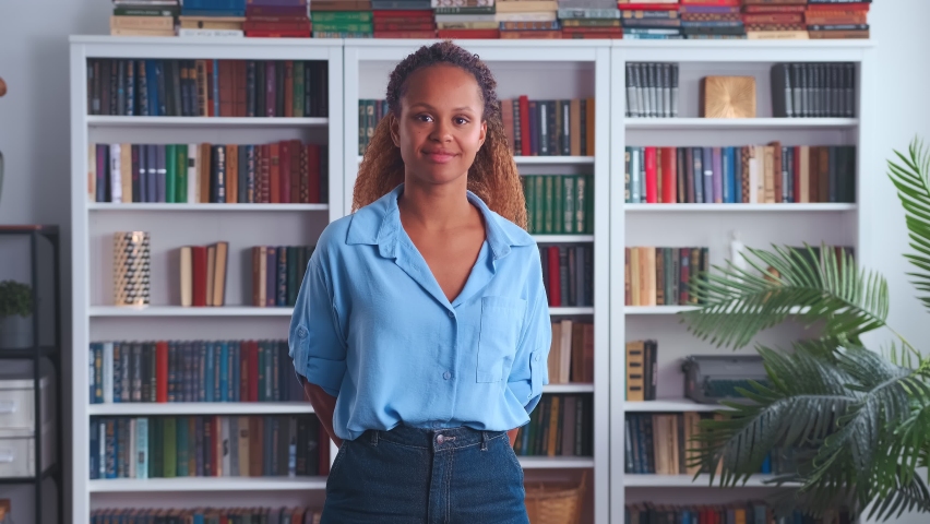 Young happy pretty African American woman student throws arms out to side and whirl with wide smile excited about going to college or receiving grant to pay for university standing in library