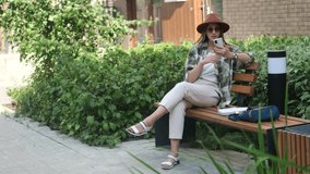 A woman is talking on a video call through a smartphone. Satisfied woman in a hat uses a smartphone while sitting on a street bench. Communications and people
