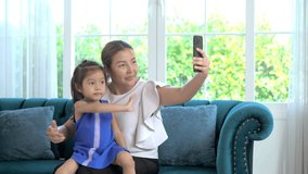 Handheld medium shot, Asian young mother and little daughter sitting on sofa enjoy to video call on smartphone in living room at home