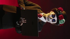 Vertical video: Goddess of death surfing internet with laptop on dios de los muertos mexican ritual, celebrating holy santa muerte with suit and hat halloween costume. Looking at website on wireless