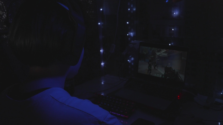 On a day off, a young woman plays a computer game sitting in the dark in front of a laptop. View from the back as a woman plays games at the computer. Hobby concept.