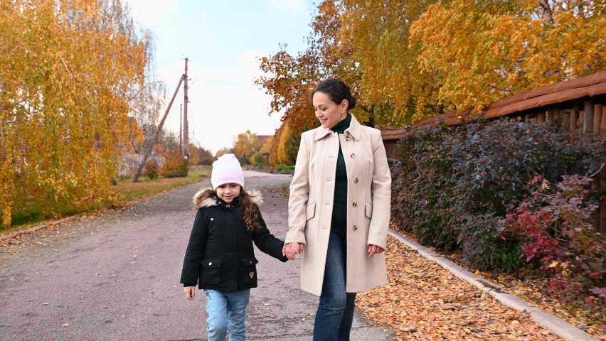 Happy young multi-ethnic woman, a loving caring mother and her lovely little daughter holding hands while walking down the street on a beautiful warm autumn day. Dry leaves fall down. Indian summer.