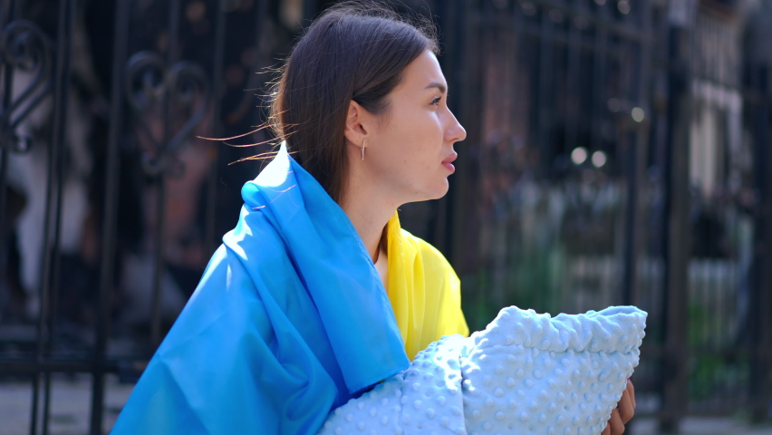 Brunette beautiful Ukrainian woman with baby wrap blanket standing in sunshine on bombed city street thinking. Side view portrait of young mother with child in ruined town outdoors
