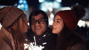 A Very Close Up Portrait of Two girls kiss a happy man with glasses on new year's eve in the middle of the street in the lights of a Christmas tree with sparklers. Concept for Christmas, New Year, Eve