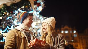 Three friends exchange gifts for New Year and Christmas. A young guy and two beautiful girls under a big Christmas glowing fir tree at a street fair. Concept for Christmas, New Year, Eve, Celebration
