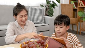 Mother feeds her grapes to son while teaching handsome kid to review lessons by tablet during the holidays, woman and son sit on warm floor and learn lesson together in living room.