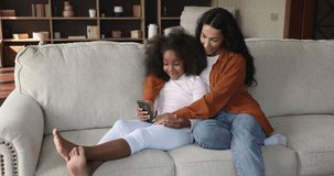African mother her cute little daughter relaxing on couch with smartphone, enjoy new cool mobile application, using gadget at home on weekend leisure. Young generation modern tech overuse, fun concept