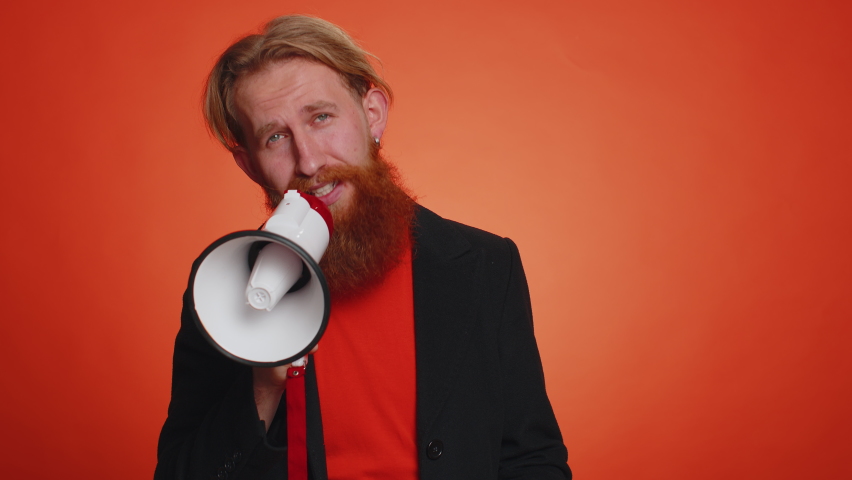 Smiling happy bearded man talking with megaphone, proclaiming news, loudly announcing sale advertisement, warning using loudspeaker to shout speech. Young redhead guy on orange studio background