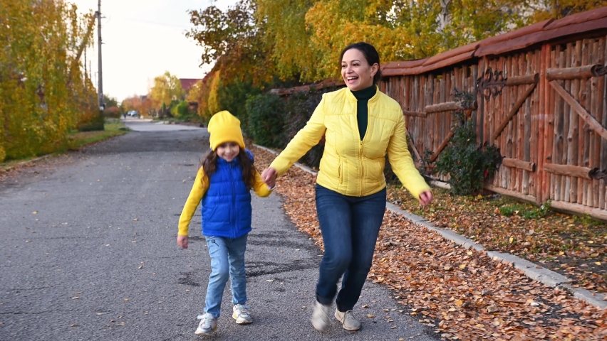 Happy family of a single mother and daughter in blue and yellow jackets, holding hands while running together along a wooden fence on a street with dried fallen leaves, in a beautiful warm autumn day | Shutterstock HD Video #1096452391