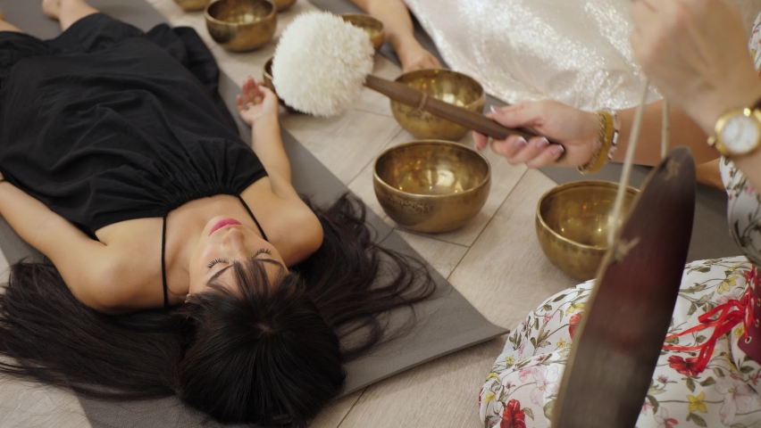 Beautiful lying woman in black having a relaxing sound therapy, specialist playing singing bowls taps the gong with soft mallet Royalty-Free Stock Footage #1096463515