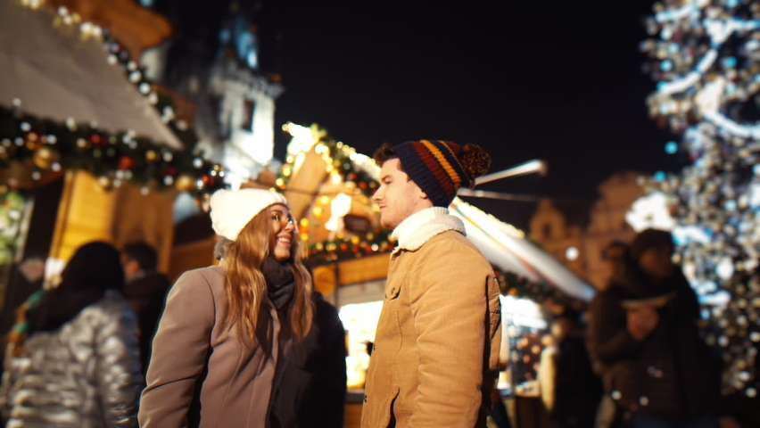 Young woman meeting up with boyfriend and kiss on christmas evening party hugging tightly together swinging under a big Christmas glowing fir tree at a street fair. Concept for Christmas, New Year