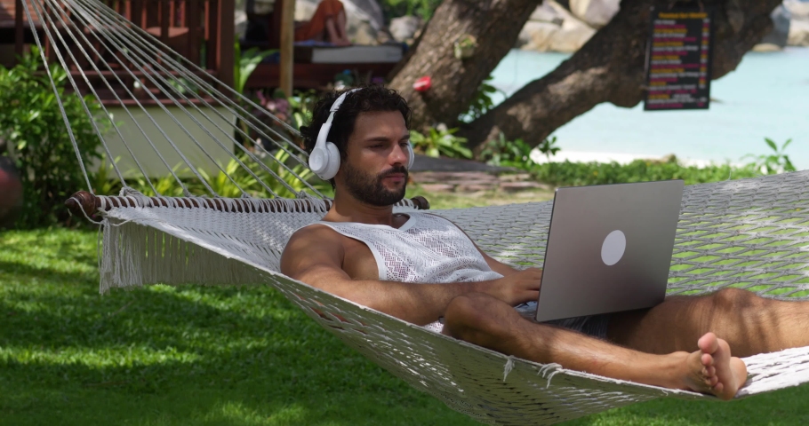 Caucasian man tourist in headphones, turn off laptop, lies in hammock, rests after work on vacation, listens music, enjoys sunbathing in coastal resort. Digital nomad Independent location entrepreneur
