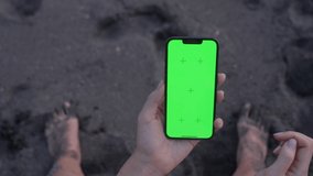 Point of View of woman holds vertical mobile and touching finger to centre touch screen sitting on beach with black sand. Use green screen for copy space closeup. Chroma key mock-up on smartphone. 
