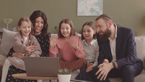 Happy Jewish couple and their three little daughters having video conversation on laptop with friends, smiling and waving at camera
