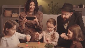 Three pretty little sisters playing with dreidels during Hanukkah. Jewish family celebrating Hanukkah together at home. Woman smiling while taking video of girls playing