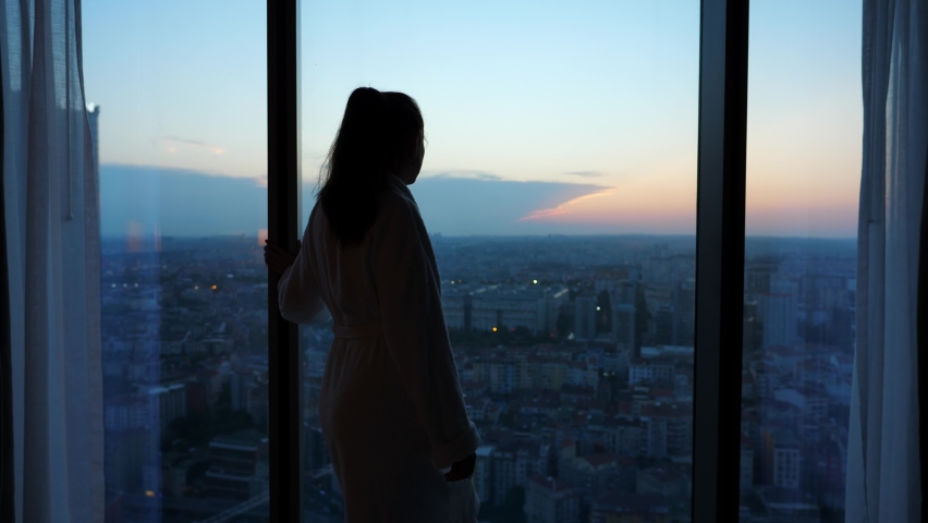 Woman is bored alone in quarantine, she stands at window and looks out at dark city. It is difficult to travel in pandemic times, it is necessary to do long quarantine upon arrival in another country Royalty-Free Stock Footage #1096473975