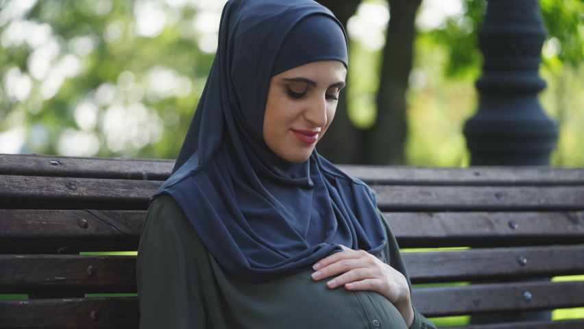 Pregnant Muslim woman in hijab sitting in park, touching belly with happy smile