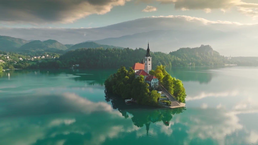 Aerial view of Church of the Assumption of Mary in the center of the lake Bled. Flying around small island on Bled Lake in Slovenia, warm morning sun and light fog