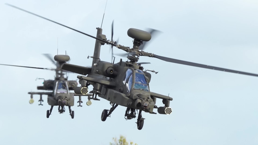 Detailed close-up of two British army Boeing Apache Attack helicopter gunships (AH64E AH-64E ArmyAir606) transition to low flight, salute from gunner, flying across frame from left to right 