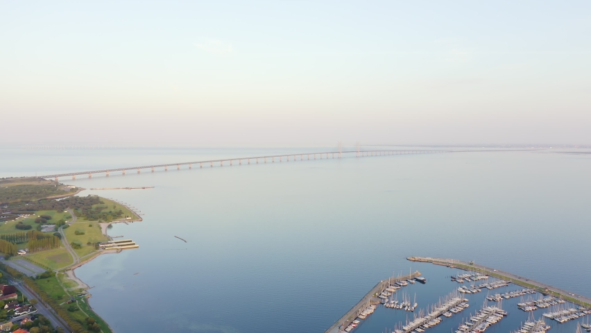 Oresund bridge. A combined road and rail cable-stayed bridge stretches across a calm strait, connecting Sweden and Denmark. The structure features tall pylons with steel cables fanning down to support the dual-level deck, carrying motorway traffic above and railway lines below. In the foreground, a marina filled with docked boats sits along a green coastline, while the bridge extends toward the horizon where it meets an artificial island that transitions into an underwater tunnel. The scene is captured in soft evening light with a clear pale sky, creating a serene and expansive atmosphere over the water. The entire link is the longest combined road and rail bridge in Europe, blending heavy infrastructure with the surrounding seascape. Hyperlapse