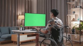 Back View Of Asian Man In Wheelchair Shaking His Head And Having A Headache While Using Mock Up Green Screen Desktop Next To The Camera At Home
