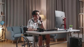 Asian Man In Wheelchair Talking On Smartphone While Editing The Video By A Desktop Next To The Camera At Home
