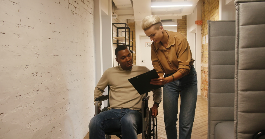 Dyed blonde woman shows black man in wheelchair project plan on clipboard in office corridor. Multinational colleagues discuss work slow motion | Shutterstock HD Video #1096488573
