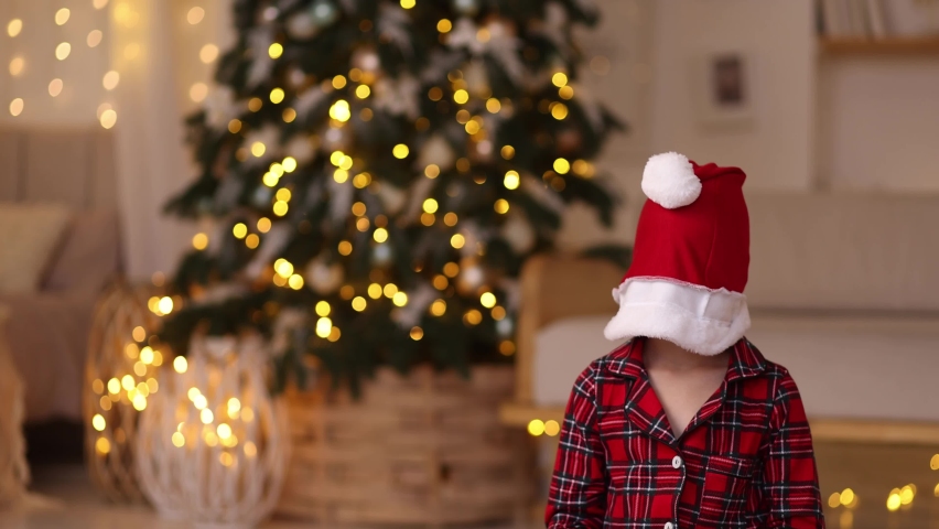 Happy girl in a red checkered New Year's pajamas sits against the background of a decorated Christmas tree joyfully looks out of a santa hat