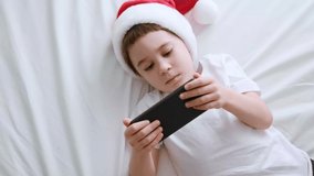 Cute 5 year boy play smartphone video game. Child in Santa Claus hat lie on bed with telephone in hand. White background. Kid use mobile phone for game or learn. Online distance e-learning. Top view.