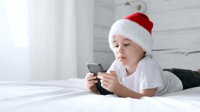 Smiling 5 year boy play smartphone video game. Said wow. Child in Santa Claus hat lie on bed. White home bedroom. Kid use mobile phone for gaming or learning. Spend time speaking with friends online.