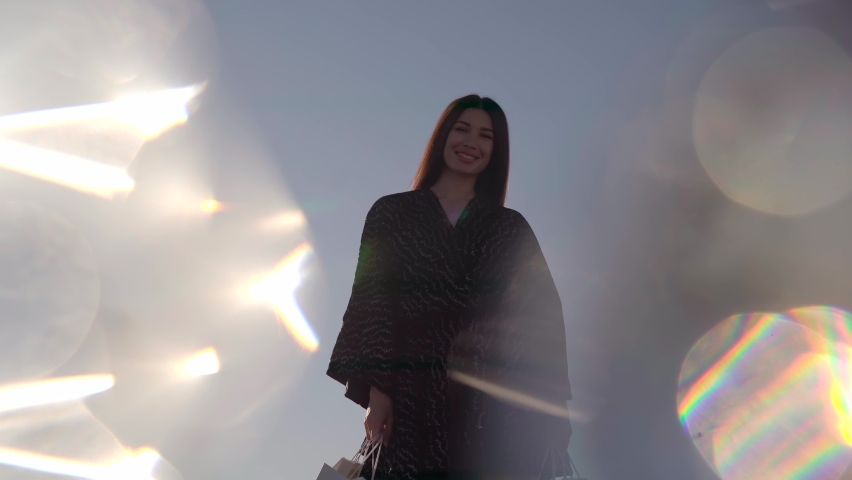 Attractive shopaholic brunette caucasian girl raising up shopping bags in hand against blue sky with flares effect. Retail, consumerism or sale concept - happy female with shopping bags. 4k video