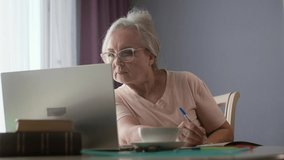 Elderly senior business woman studying a new job remotely on a laptop taking notes in a notebook. Retired looking for a job.