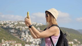 young woman traveler in black glasses and panama hat on background of town makes photos and records videos. Girl tourist blogger make overview of journey while standing on top of mountain above city.